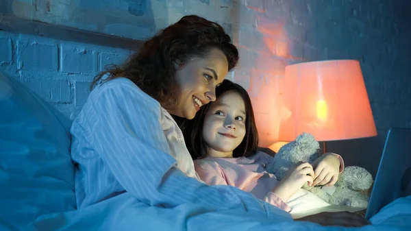 Улыбающийся ребенок с плюшевым мишкой смотрит на мать с помощью ноутбука на кровати — стоковое фото