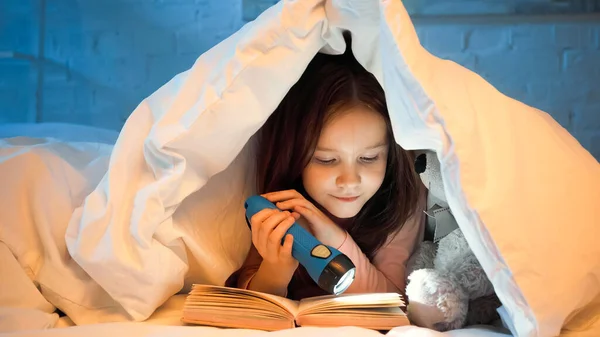 Preteen girl with flashlight reading book on bed in evening — Stock Photo
