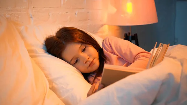 Kid leitura livro perto de cobertor borrado na cama — Fotografia de Stock