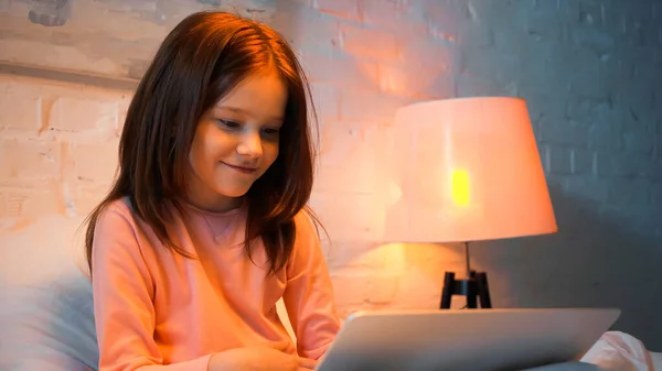 Lächelndes Kind mit Laptop auf verschwommenem Vordergrund im Schlafzimmer während der Nacht — Stockfoto