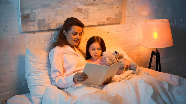 Mãe sorridente no pijama livro de leitura perto da criança com brinquedo macio na cama — Fotografia de Stock