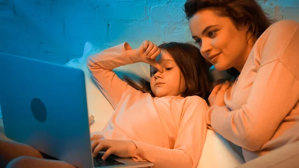 Mulher sorridente deitada perto da filha usando laptop desfocado na cama — Fotografia de Stock