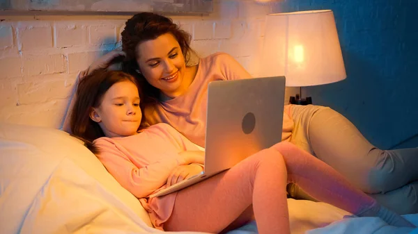 Kid with laptop lying near positive mom on bed — Stock Photo