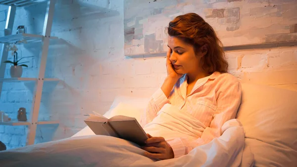 Mujer agotada con libro sentado en la cama por la noche - foto de stock