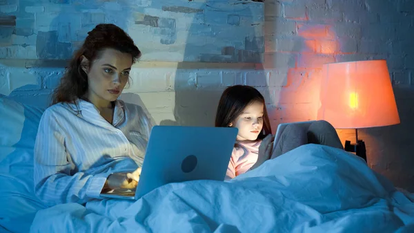 Freelancer using laptop near daughter with digital tablet on bed — Stock Photo