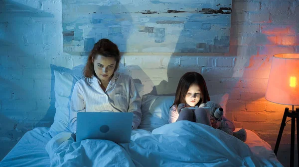 Mother and daughter with soft toy using gadgets on bed in night — Stock Photo