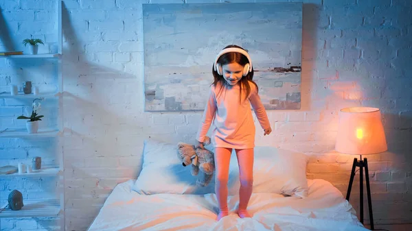 Alegre niño en auriculares sosteniendo juguete suave en la cama - foto de stock