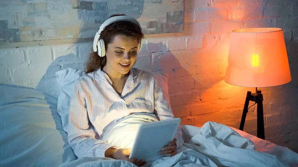 Mujer sonriente usando tableta digital y auriculares en el dormitorio - foto de stock