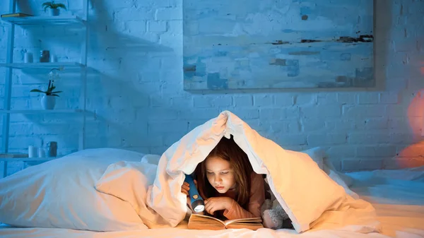 Child under blanker reading book and holding flashlight — Stock Photo