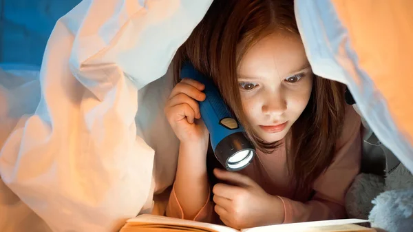 Kid holding flashlight and reading book under blanket in bedroom — Stock Photo