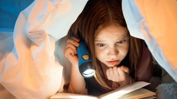 Niño concentrado con linterna libro de lectura en la cama - foto de stock