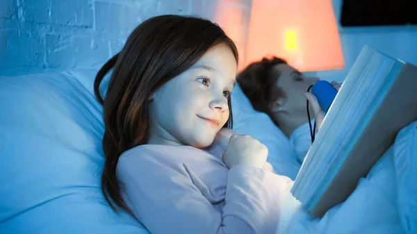 Fille souriante avec lampe de poche lecture livre près de mère endormie sur fond flou — Photo de stock