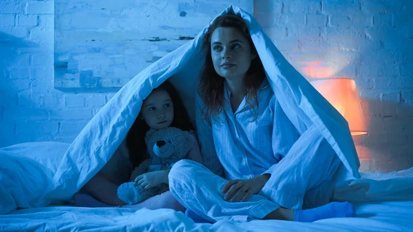 Madre e hijo con juguete suave sentado debajo de la manta en la cama durante la noche - foto de stock