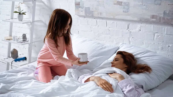 Улыбающаяся девушка держит чашку кофе рядом с матерью на кровати — стоковое фото