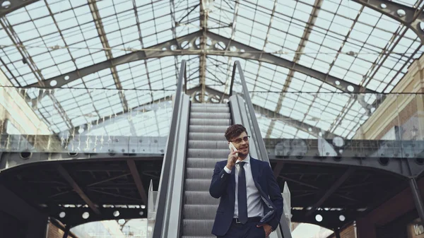 Hombre de negocios feliz en gafas hablando en el teléfono inteligente y de pie con la mano en el bolsillo en las escaleras mecánicas - foto de stock