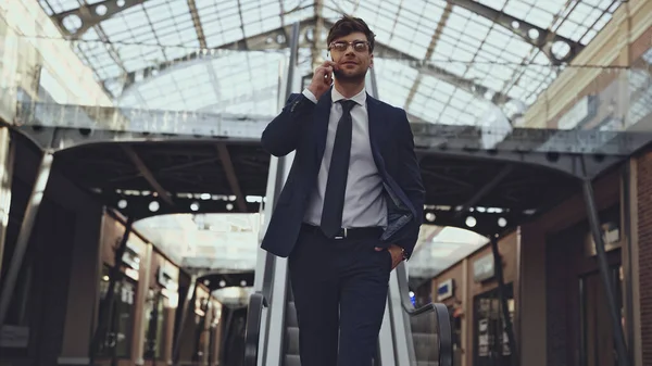 Homem de negócios feliz em óculos falando no smartphone e andando com a mão no bolso no centro comercial — Fotografia de Stock