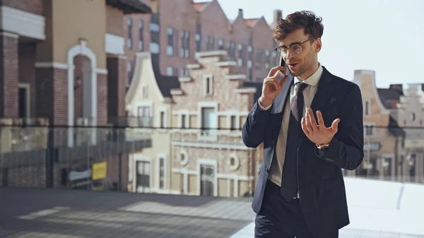 Cheerful businessman in glasses and suit talking on smartphone in modern shopping center — Stock Photo