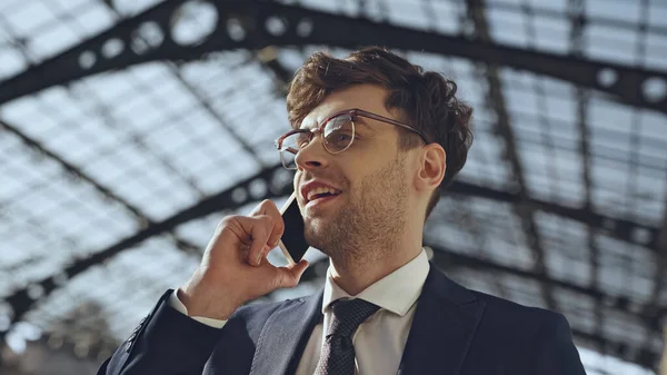Vista de ángulo bajo de hombre de negocios alegre en gafas y traje hablando en el teléfono inteligente en el centro comercial moderno - foto de stock