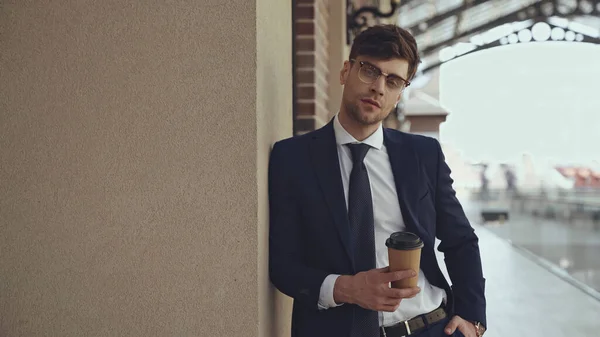 Бизнесмен в очках и костюме держит кофе, чтобы пойти и позировать в торговом центре — стоковое фото