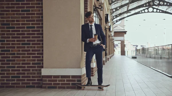 Longitud completa de hombre de negocios en gafas y traje sosteniendo café para ir posando cerca de penny board en el centro comercial - foto de stock