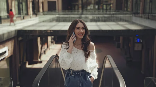 Cheerful young woman talking on smartphone while standing with hand in pocket near escalator — Stock Photo