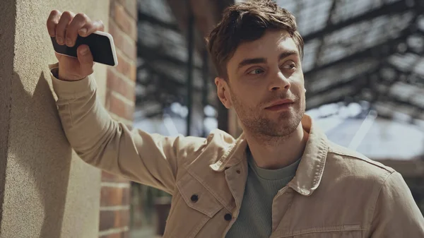 Good-looking man in beige jacket holding smartphone and looking away in shopping mall — Stock Photo