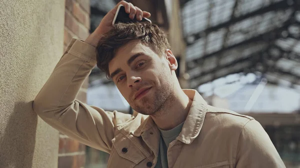 Hombre guapo en chaqueta beige sosteniendo el teléfono inteligente y mirando a la cámara en el centro comercial - foto de stock