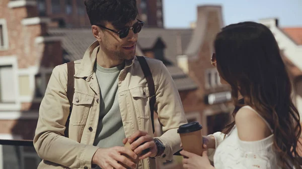 Casal elegante em óculos de sol segurando copos de papel e falando fora — Fotografia de Stock