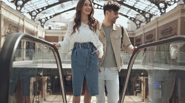 Счастливая женщина позирует с рукой в кармане рядом с мужчиной на эскалаторе в торговом центре — стоковое фото