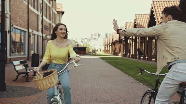 Homem feliz acenando mão perto de mulher andar de bicicleta na rua — Fotografia de Stock