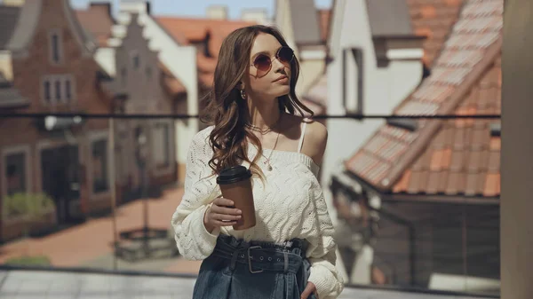 Стильная молодая женщина в солнечных очках держит кофе, чтобы выйти на улицу — стоковое фото