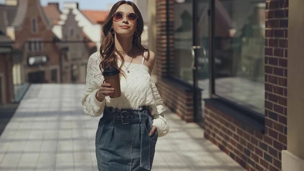 Stylish young woman in sunglasses holding coffee to go and walking outside — Photo de stock