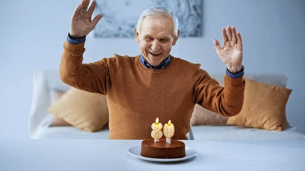 Jubiloso anciano celebrando cumpleaños delante de la torta con velas encendidas en la sala de estar - foto de stock