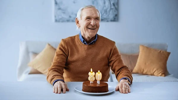 Feliz anciano celebrando cumpleaños delante de la torta con velas encendidas en la sala de estar en la sala de estar - foto de stock