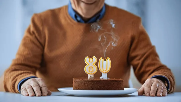 Vista recortada del anciano celebrando su cumpleaños frente a la torta con velas apagadas sobre fondo gris - foto de stock