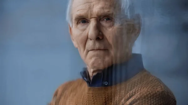 Portrait of elderly man looking at camera through rainy window on grey background behind rainy glass — Stock Photo