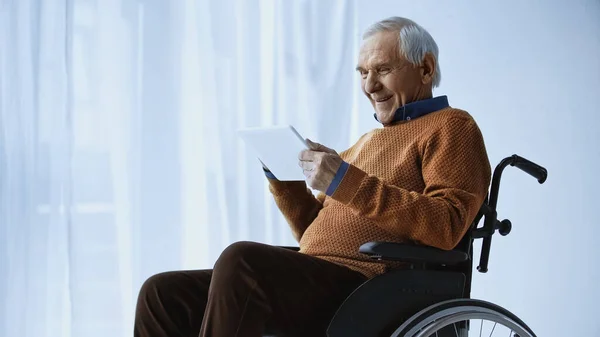 Lächelnder älterer Mann im Rollstuhl schaut im Altenheim auf Tablet in der Hand — Stockfoto