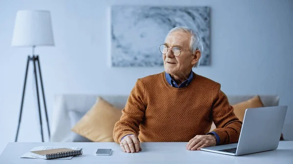 Homme âgé assis à table avec ordinateur portable et smartphone à la maison — Photo de stock