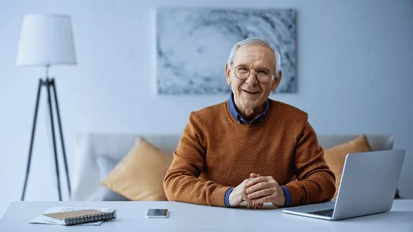 Счастливый пожилой человек сидит с сжатыми руками за столом с ноутбуком и сотовым телефоном в гостиной — стоковое фото