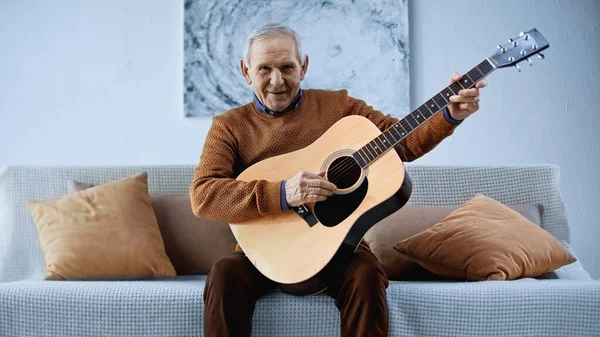 Anciano sonriente sentado en un sofá con guitarra en la sala de estar - foto de stock