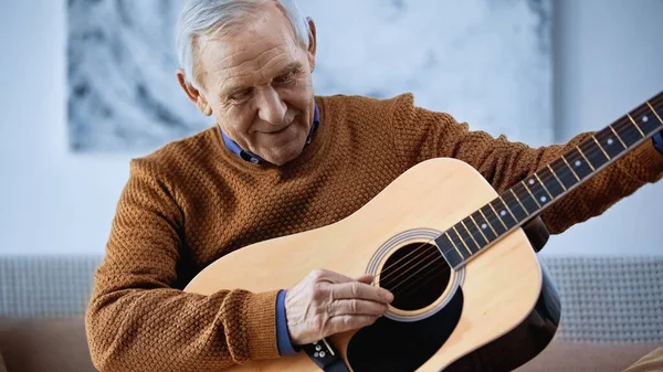 Усміхнений літній чоловік сидить на дивані і грає на гітарі у вітальні — стокове фото