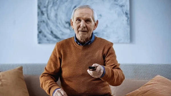 Homme âgé concentré assis sur le canapé avec télécommande tv et regardant la caméra à la maison — Photo de stock