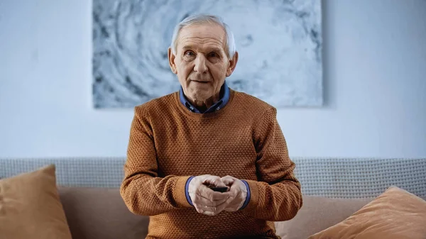 Uomo anziano seduto sul divano con telecomando TV e guardando la fotocamera a casa — Foto stock