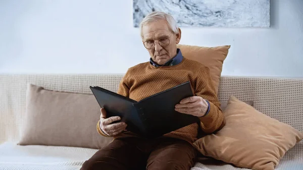 Homem idoso sentado no sofá com álbum de família em casa — Fotografia de Stock