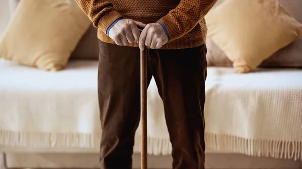 Обрезанный вид пожилой человек с тростью стоя возле дивана дома — стоковое фото