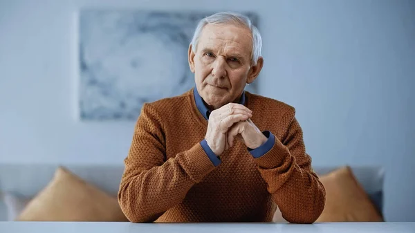 Ernster älterer Mann mit geballter Hand in der Nähe des Gesichts, der zu Hause am Tisch sitzt — Stockfoto