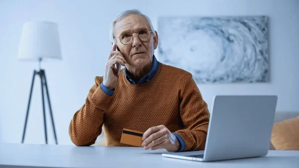 Grave uomo anziano seduto con carta di credito vicino al computer portatile e parlando sul cellulare a casa — Foto stock