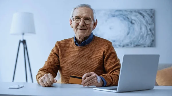 Homem idoso feliz em óculos sentado perto de laptop e celular e segurando cartão de crédito na sala de estar — Fotografia de Stock