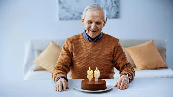 Счастливый пожилой человек празднует день рождения перед тортом с горящими свечами в гостиной — стоковое фото