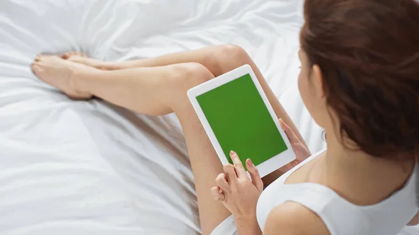 Vista ad alto angolo della giovane donna riccia seduta sul letto e utilizzando tablet digitale con schermo verde — Foto stock